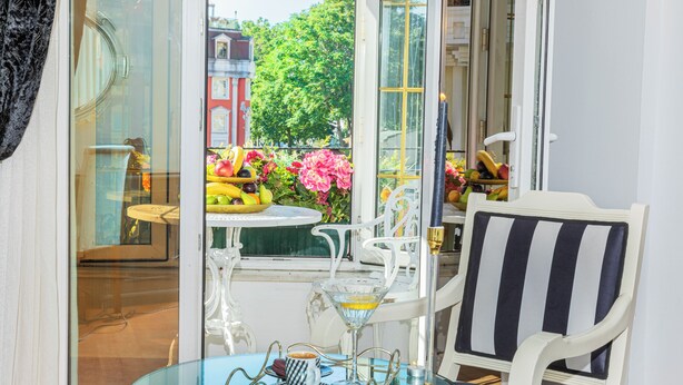Et værelse med et glastilbørsbord, hvide stole, en stribet stol og udsigt til en balkon med blomster og frugt.