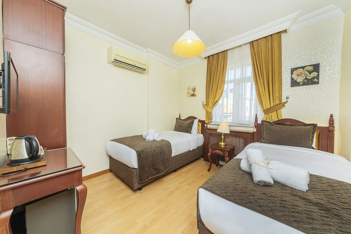 A hotel room with two beds, a wooden desk with a kettle, a small table with a lamp, and a window with curtains.