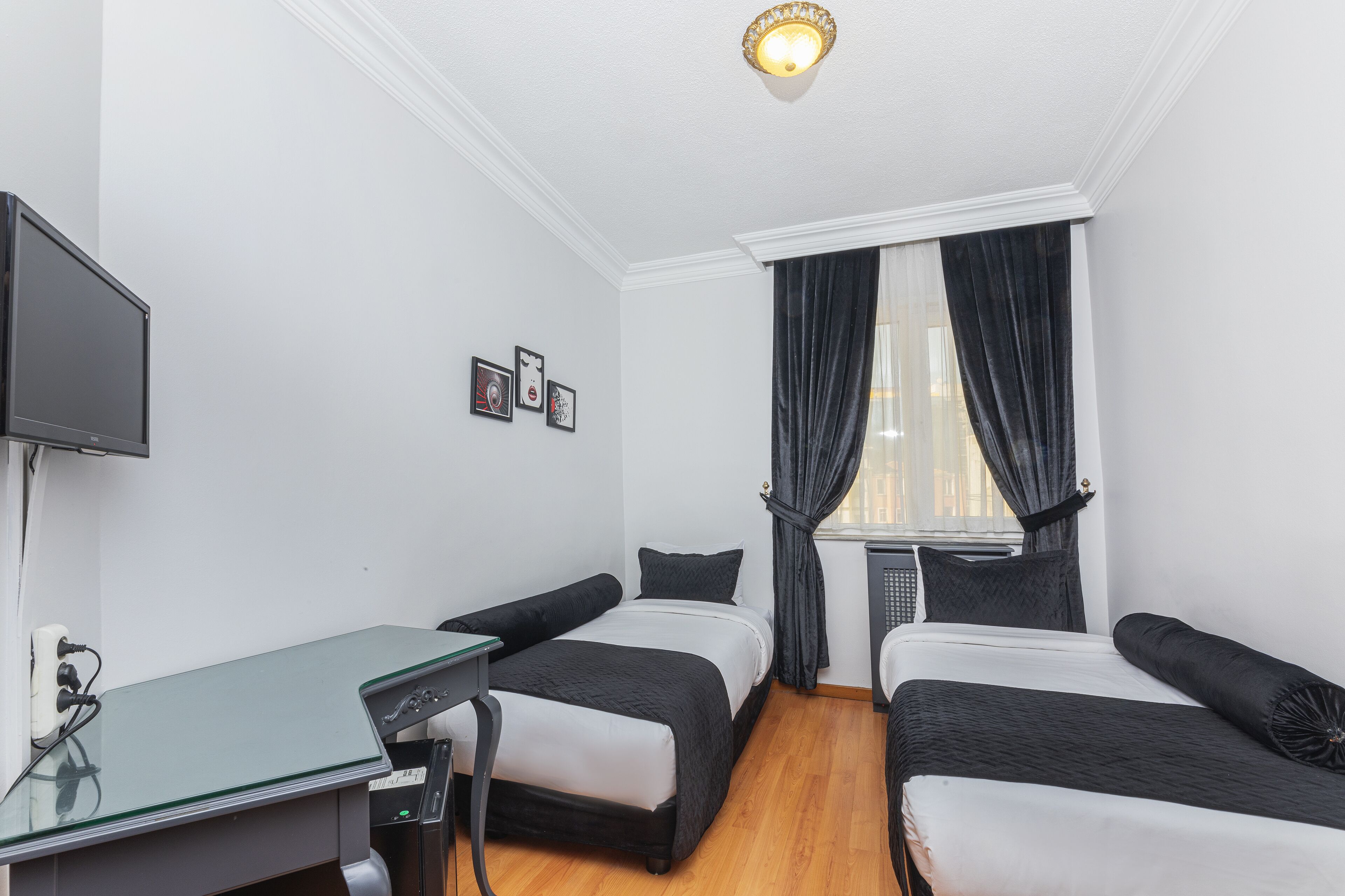 A hotel room with two single beds, a desk with a keyboard, a flat-screen TV mounted on the wall, and a window with dark curtains.