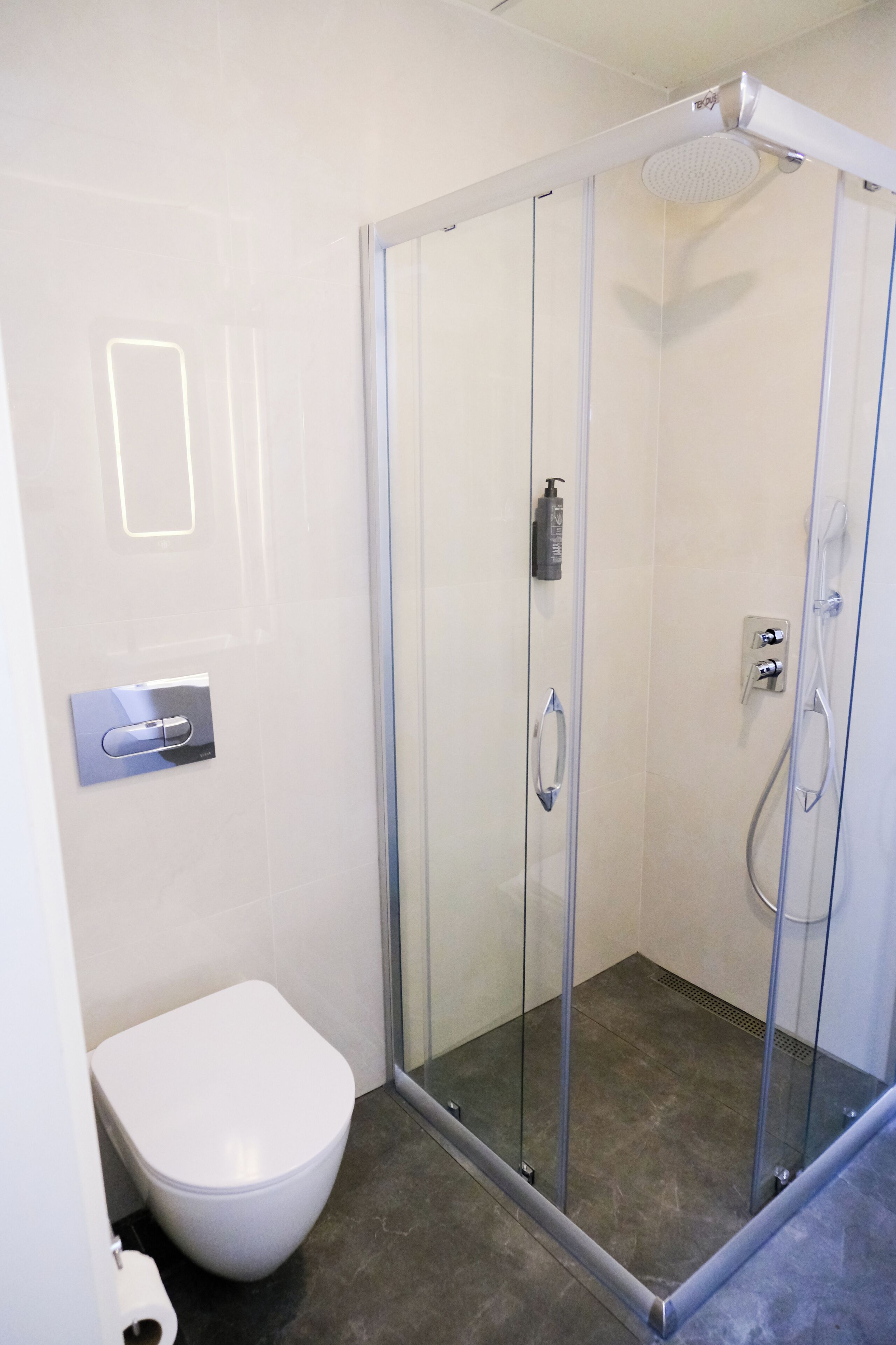 Un bany modern amb dutxa amb envàs de vidre, vàter blanc i dosificador de sabó fixat a la paret.