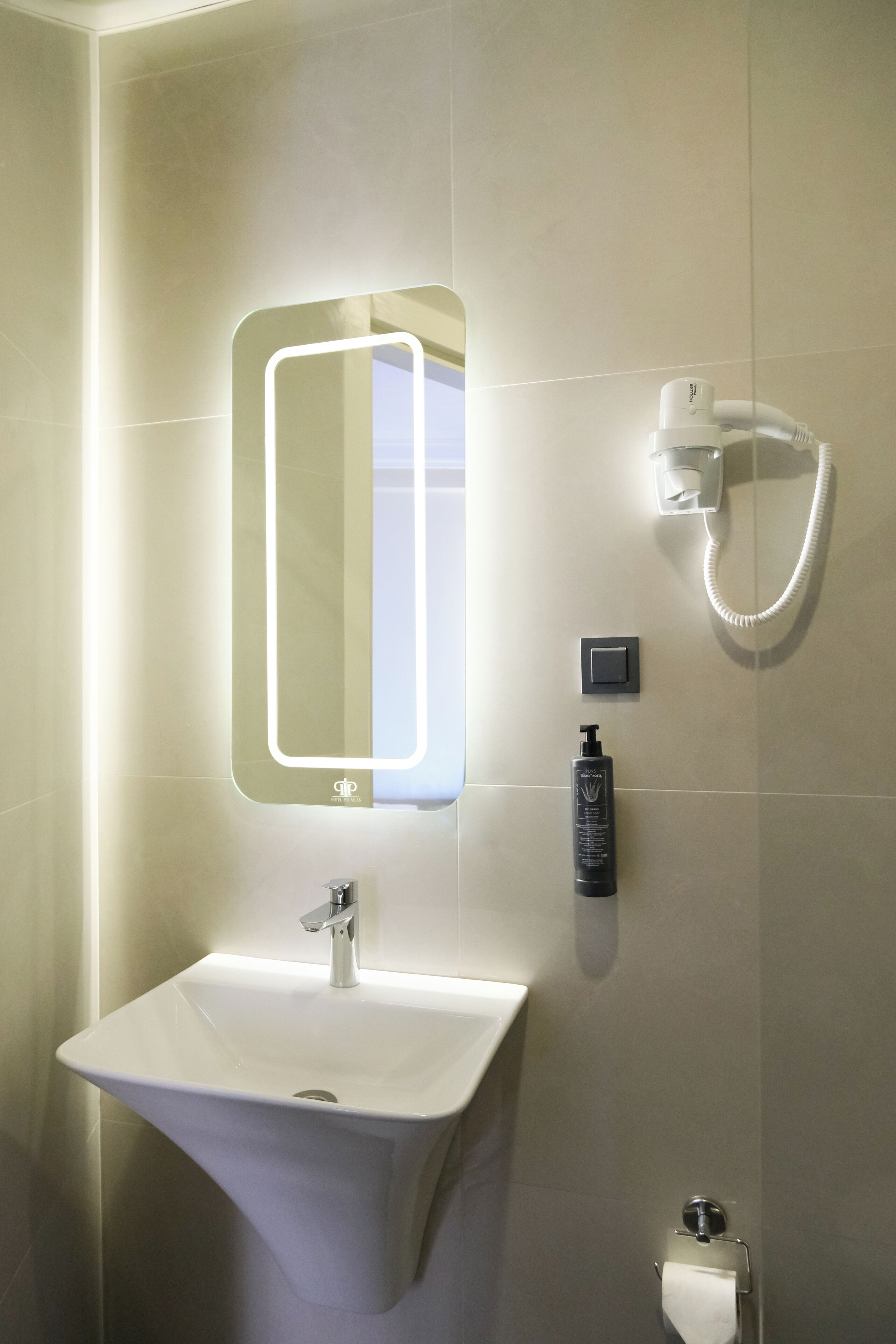 A bathroom with a white sink, a mirror with integrated lighting, a handheld showerhead, and a soap dispenser.