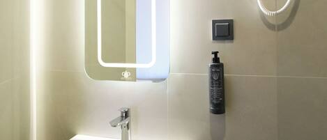 A bathroom with a white sink, a mirror with integrated lighting, a handheld showerhead, and a soap dispenser.