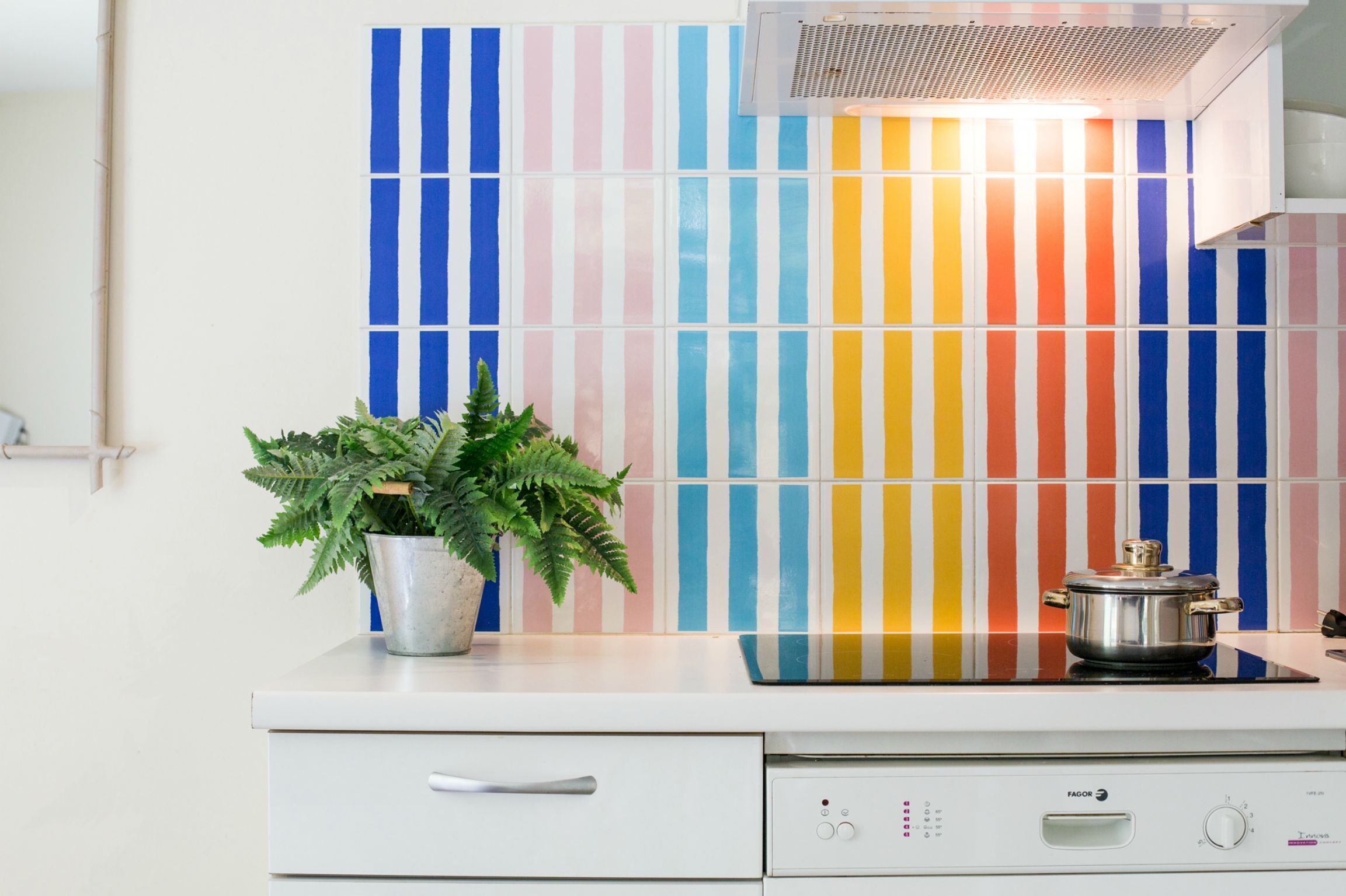 A kitchen with a white countertop, a stainless steel pot, a potted plant, and a wall with colorful vertical stripes.