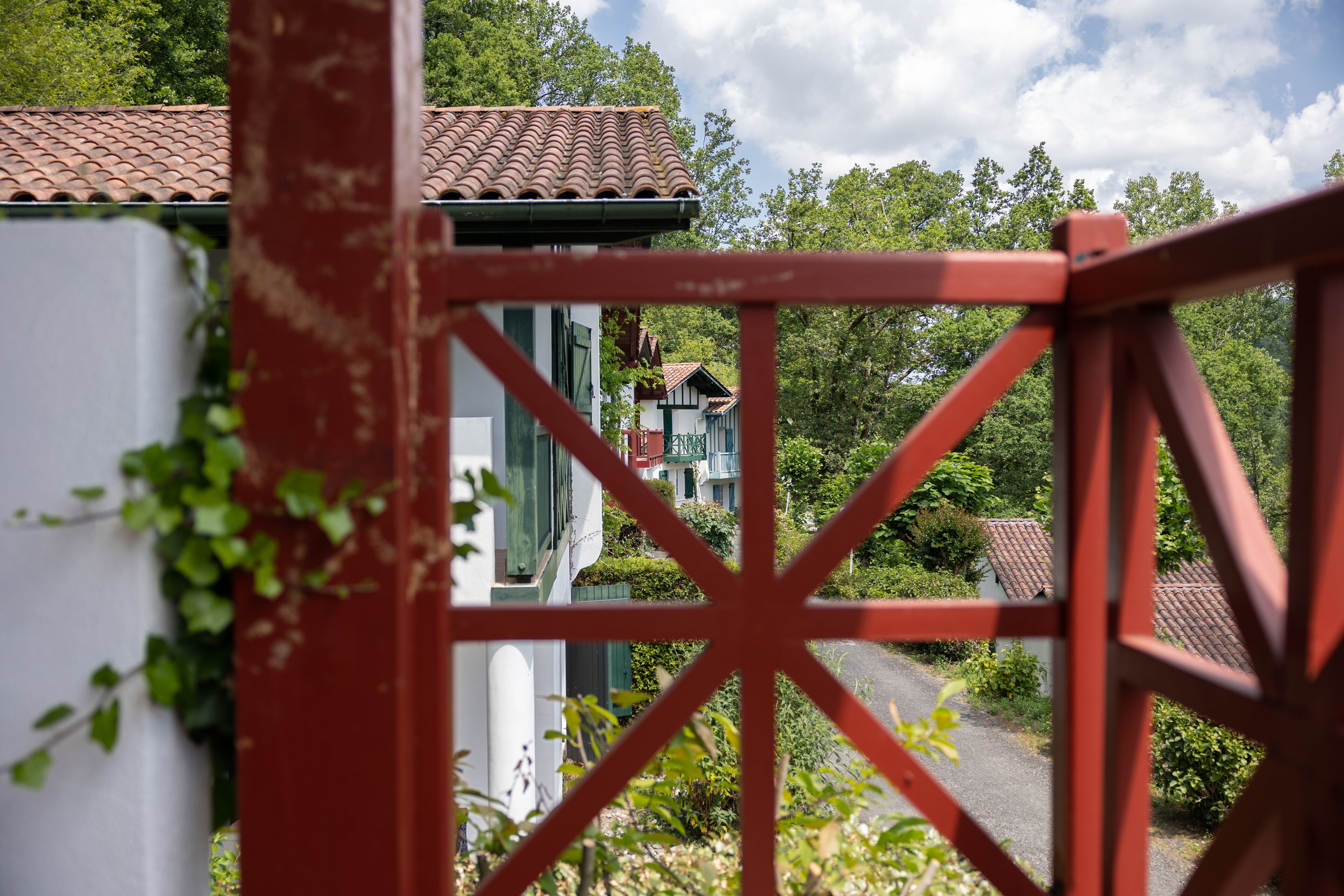 Un cancello di metallo rosso che offre una vista su un edificio con tetto in tegole e vegetazione sullo sfondo.