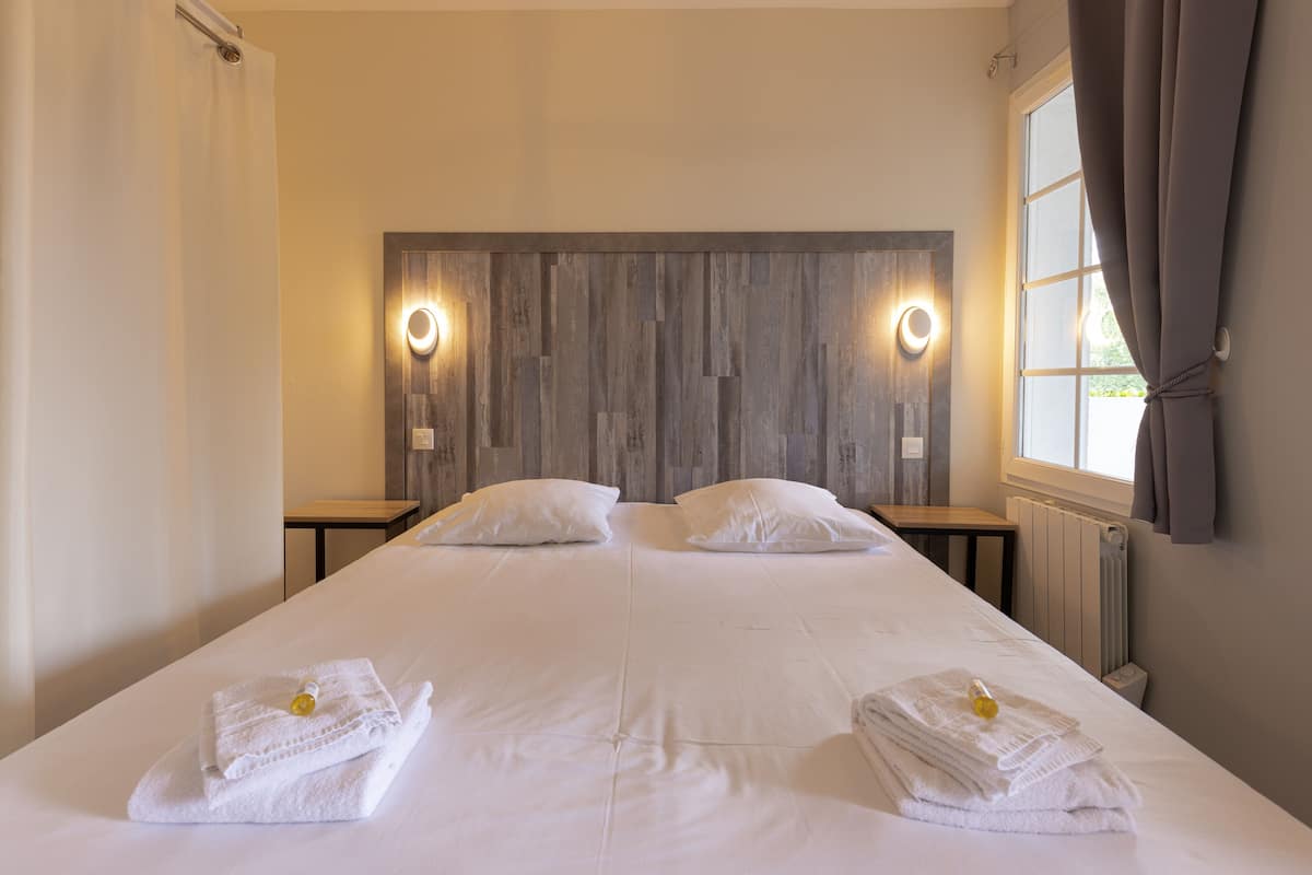 Una cama doble con sábanas blancas, dos mesitas de noche, un cabecero de madera y una ventana con cortinas.
