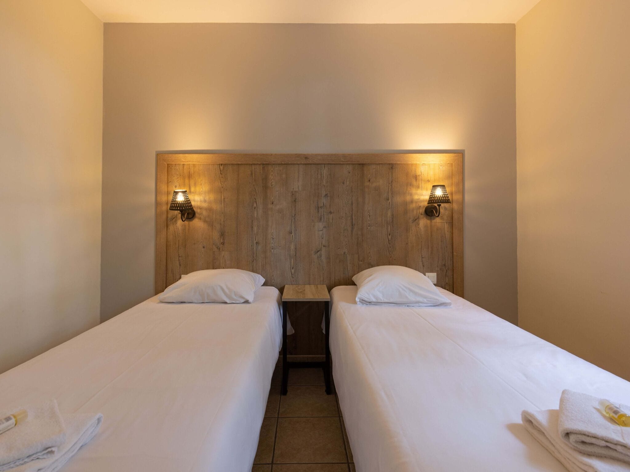 A hotel room with two single beds, each with a pillow and a neatly folded towel. There are two wall-mounted lamps on either side of the headboard.