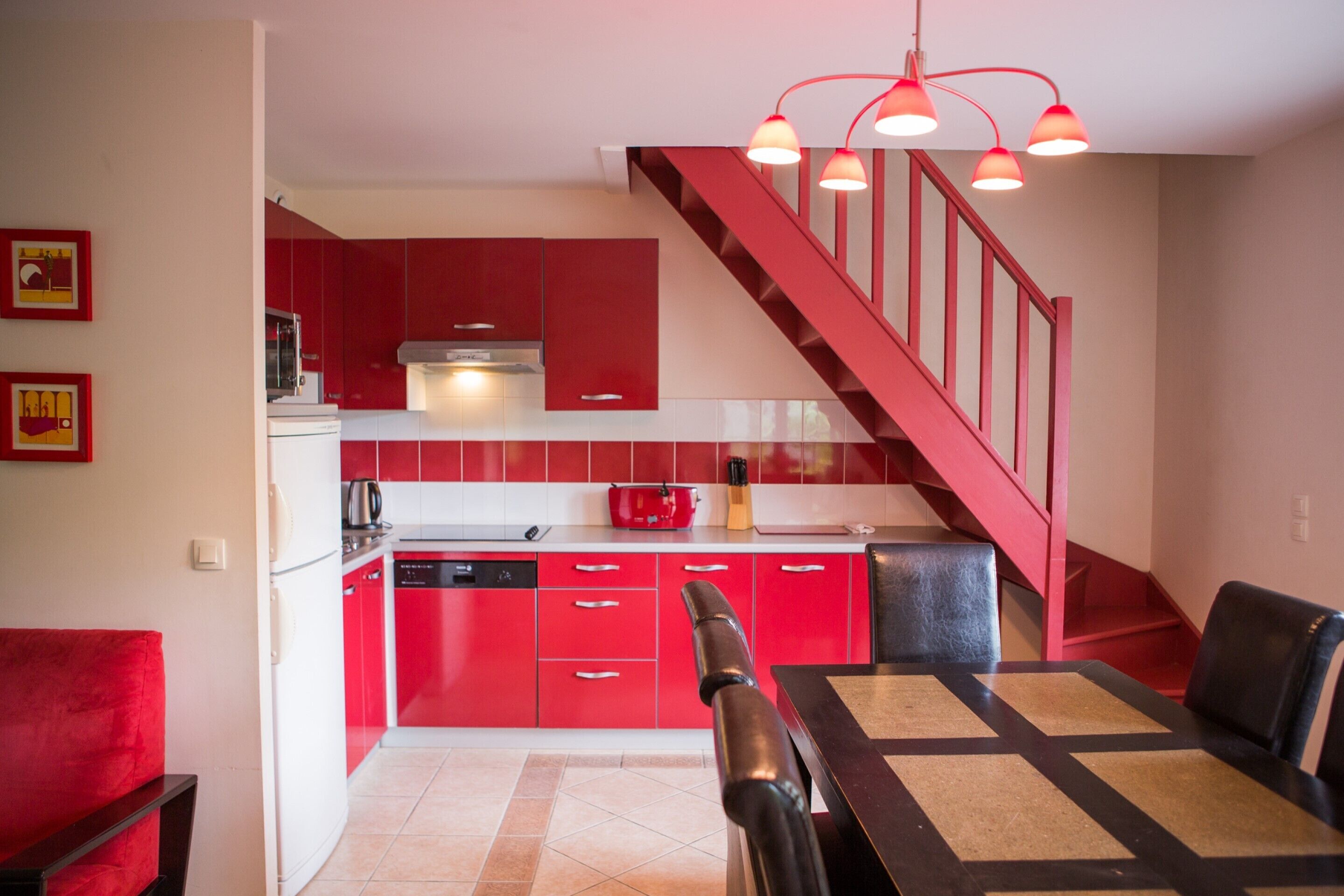 A modern kitchen with red cabinets, a dining area with a table and chairs, and a red staircase.