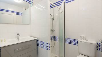Un baño con lavamanos blanco, espejo, ducha con mampara de vidrio y retrete.
