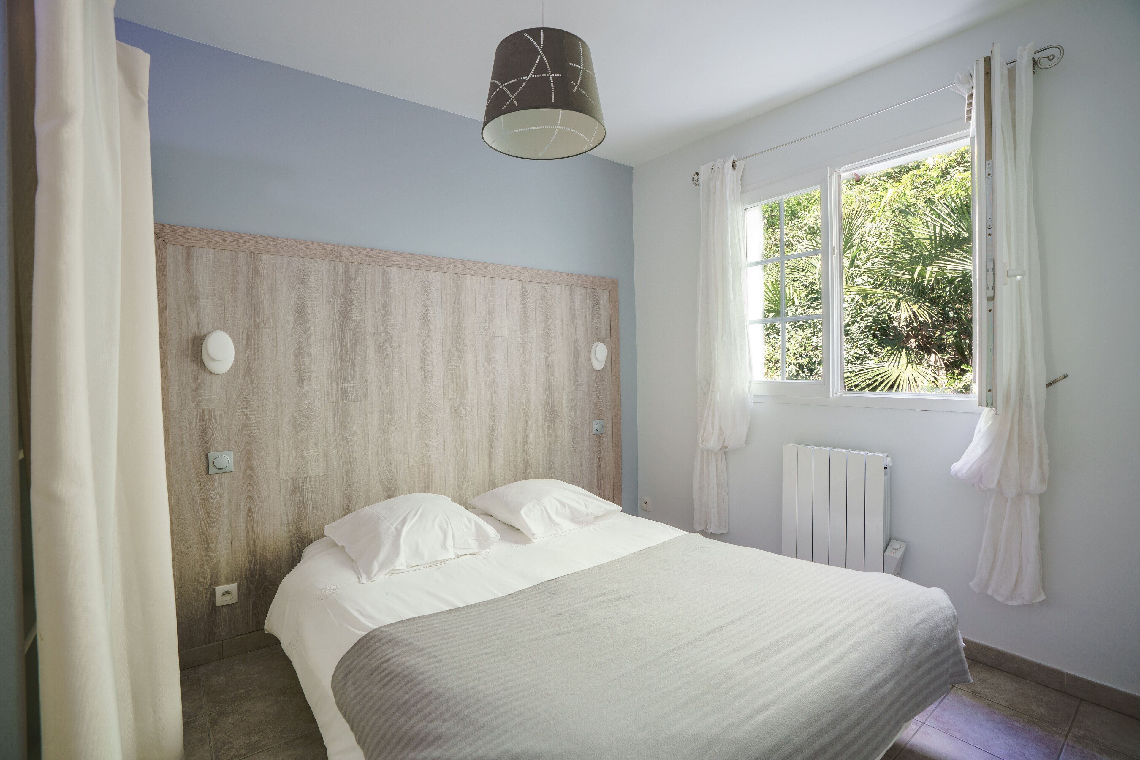 A bedroom with a bed, a window with curtains, a wooden headboard, and a ceiling light.