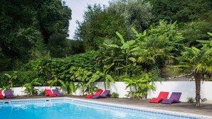 A swimming pool with red and purple lounge chairs, surrounded by lush greenery and a white wall. - Les Collines Iduki (La Bastide-Clairence)