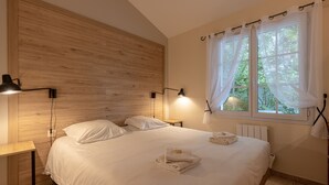 A neatly made bed with white linens, two bedside lamps, a wooden headboard, a window with sheer curtains, and a radiator. - Les Collines Iduki (La Bastide-Clairence)