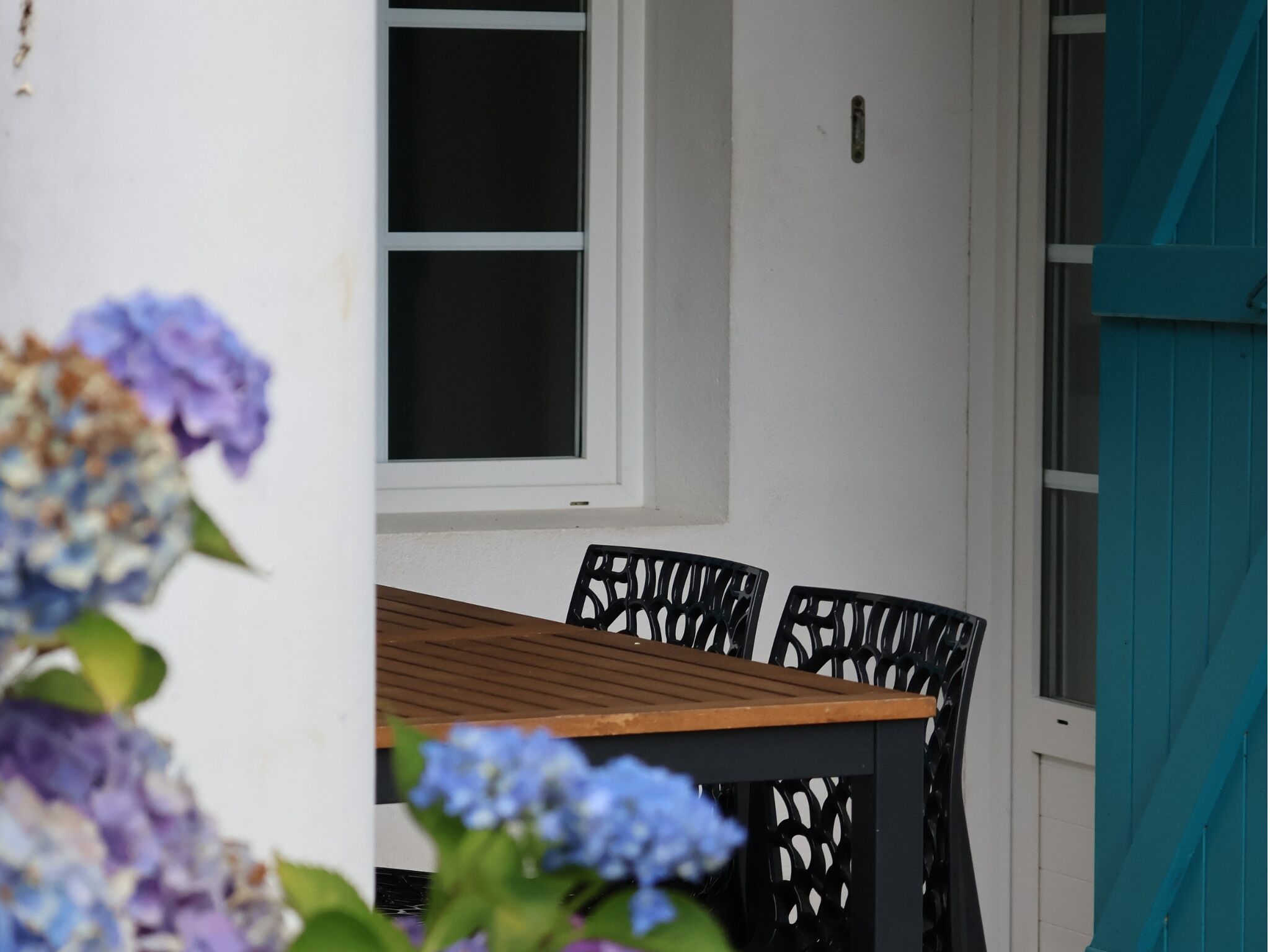 Una mesa de madera con cuatro sillas, un ventanal y una puerta con contraventanas color turquesa.