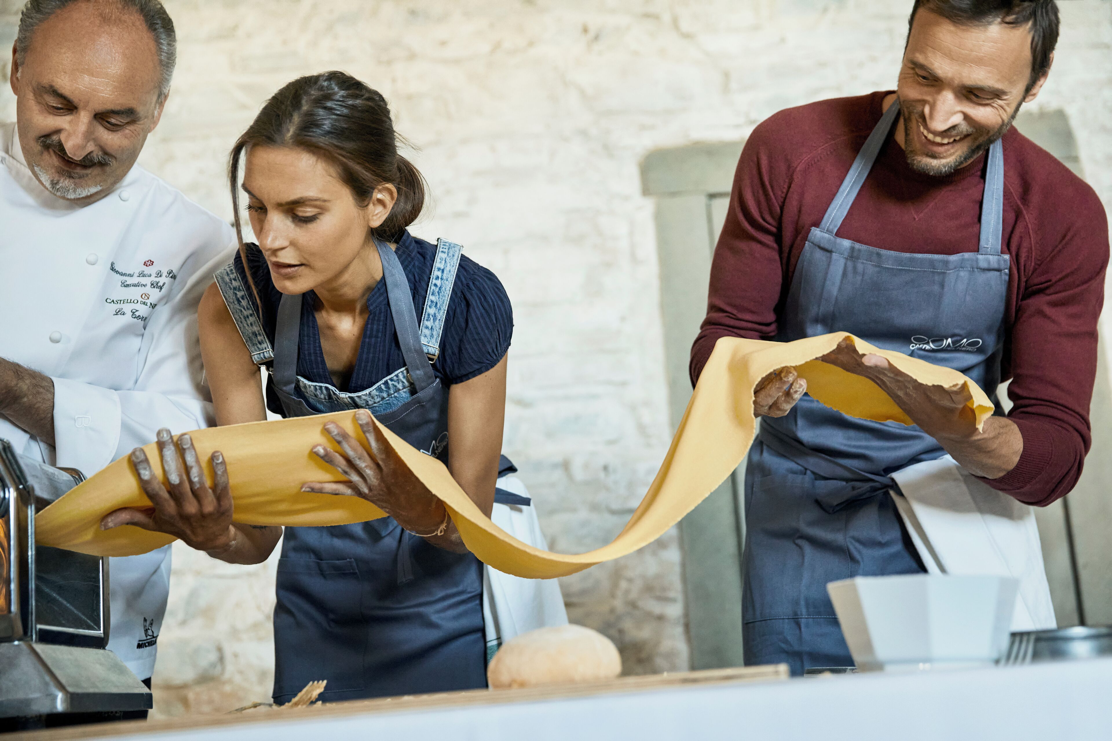 Three individuals in a kitchen setting, one wearing a chef's uniform, another in a casual shirt with an apron, and the third in a maroon shirt with an apron. They are handling a large sheet of yellow material, possibly dough or pastry.