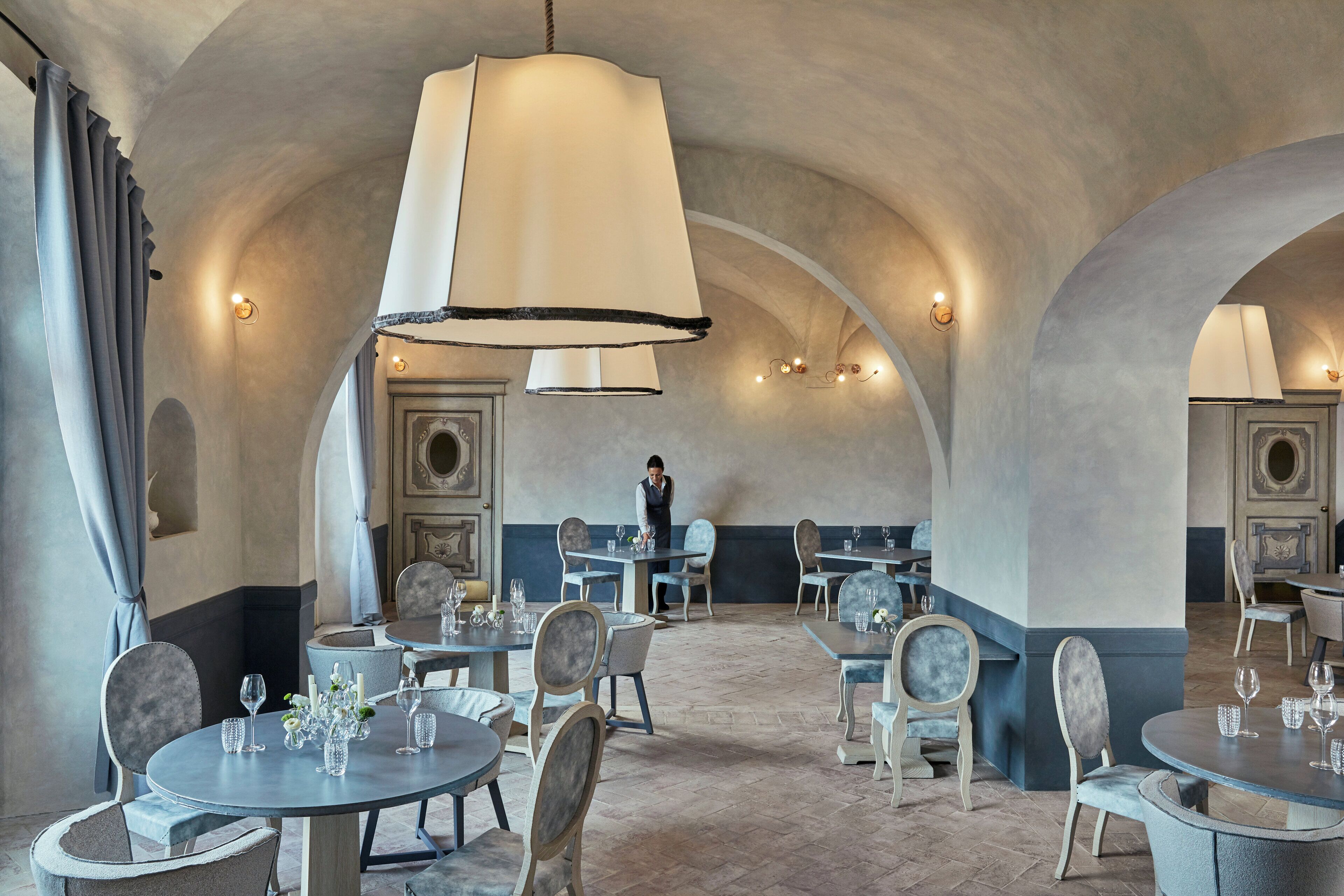 Un restaurant avec des tables et des chaises rondes, un serveur et un plafond haut orné d’arcs.