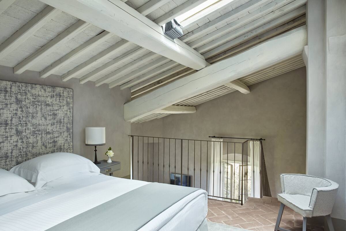 Un dormitorio con techo inclinado, una cama con ropa blanca, una mesita de noche con lámpara y una silla.