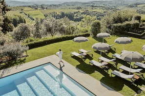 A poolside area with lounge chairs and umbrellas, a person walking, and a view of the countryside. - COMO Castello Del Nero (Barberino Tavarnelle)