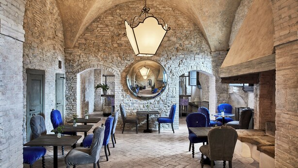 A spacious interior with high arched ceilings, stone walls, and a central round mirror reflecting the room. Tables and chairs are arranged for dining or socializing. - COMO Castello Del Nero (Barberino Tavarnelle)