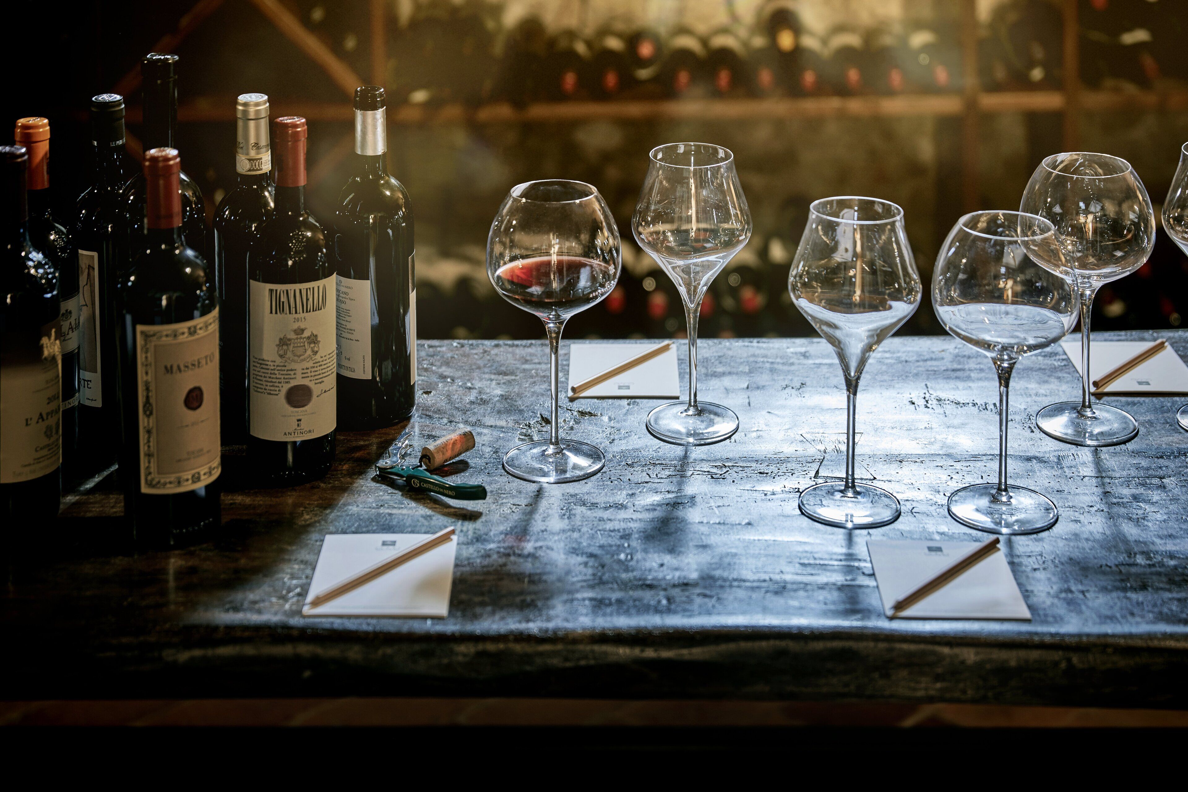 Varias botellas de vino y copas sobre una mesa de madera, con estanterías llenas de vino al fondo.