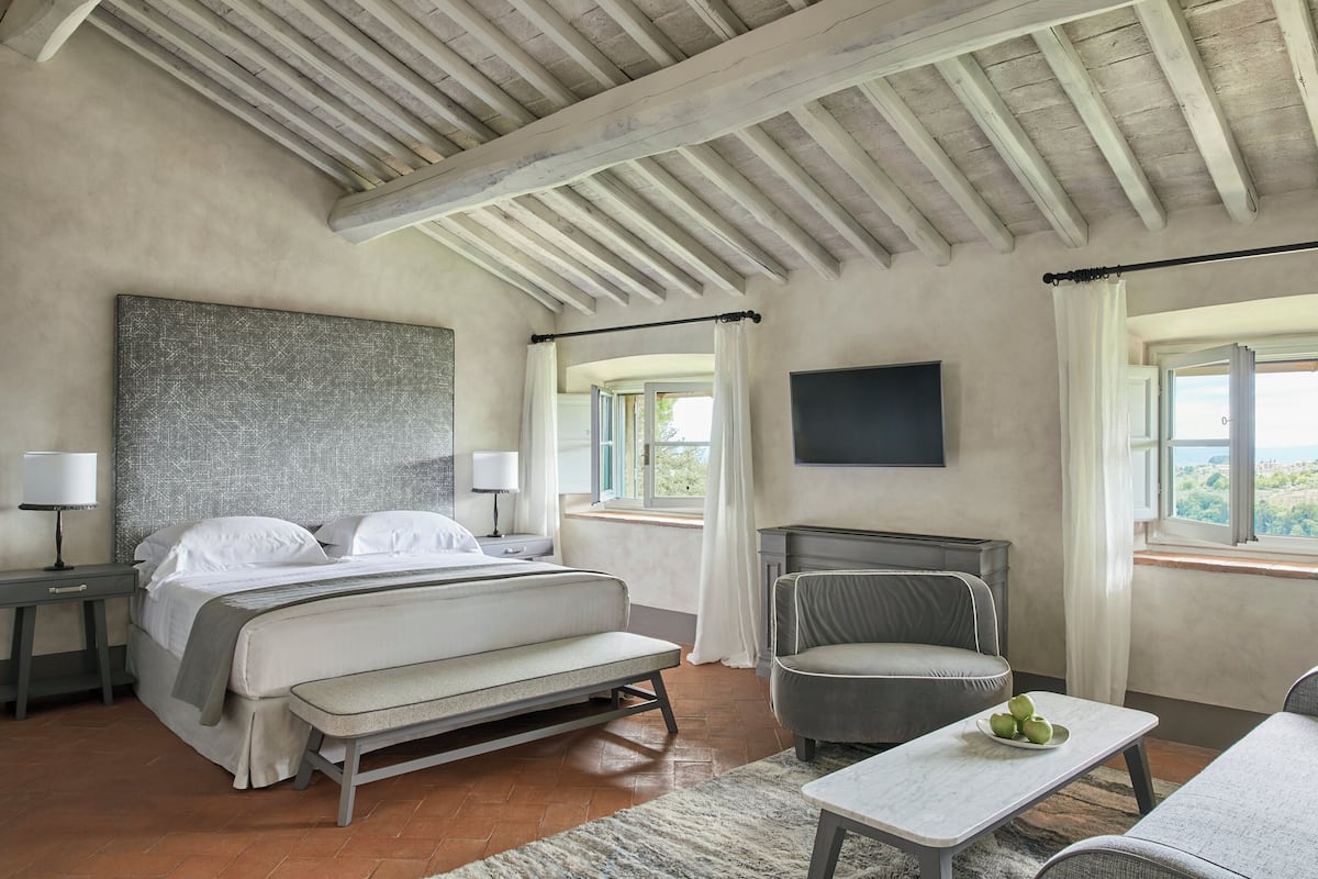 Un dormitorio amplio con una cama grande, una mesita de noche con lámpara, una silla, una pequeña mesa con fruta y un televisor montado en la pared.