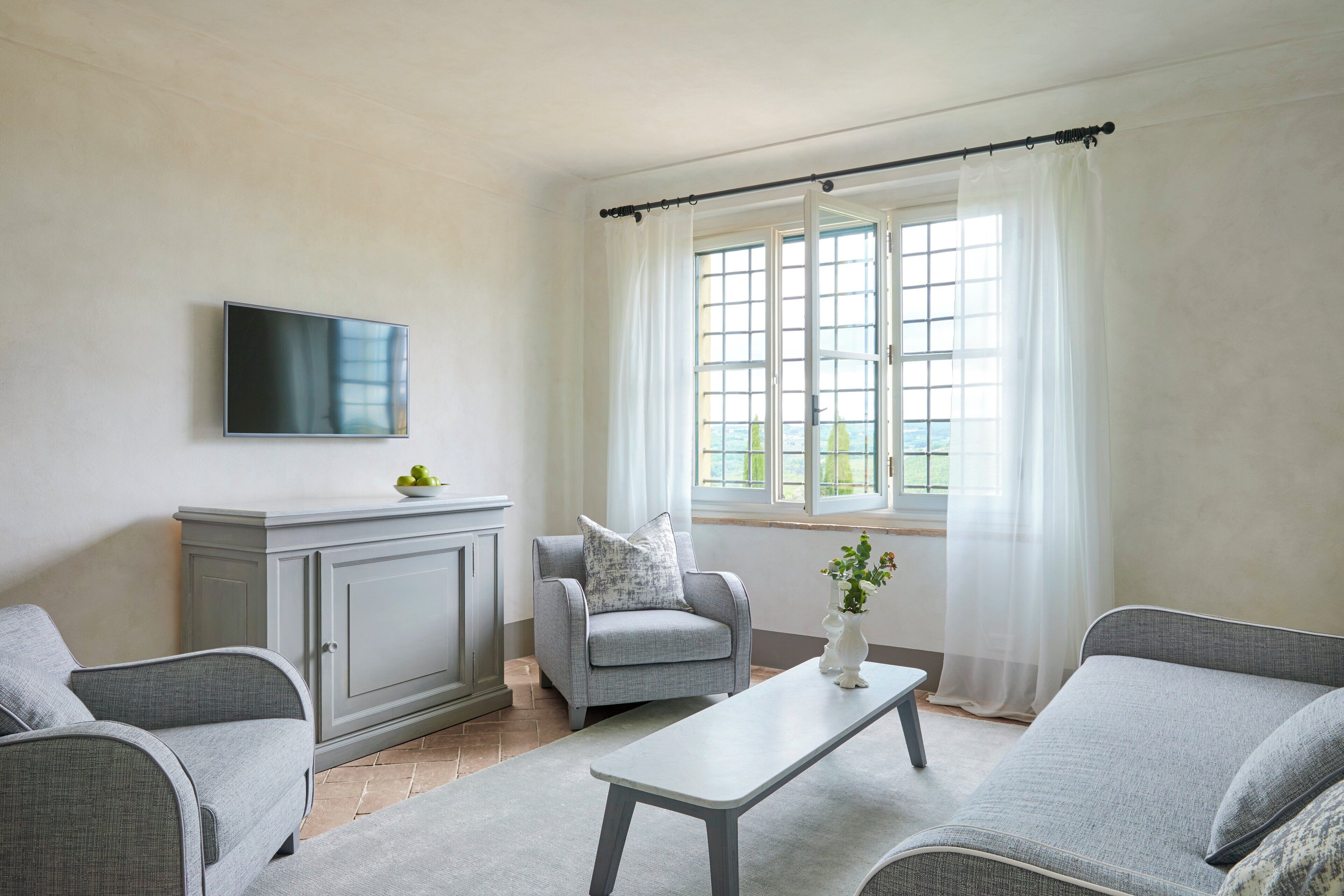A living room with grey sofas, a flat-screen TV, a grey cabinet, and a window with white curtains.