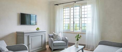 A living room with grey sofas, a flat-screen TV, a grey cabinet, and a window with white curtains.