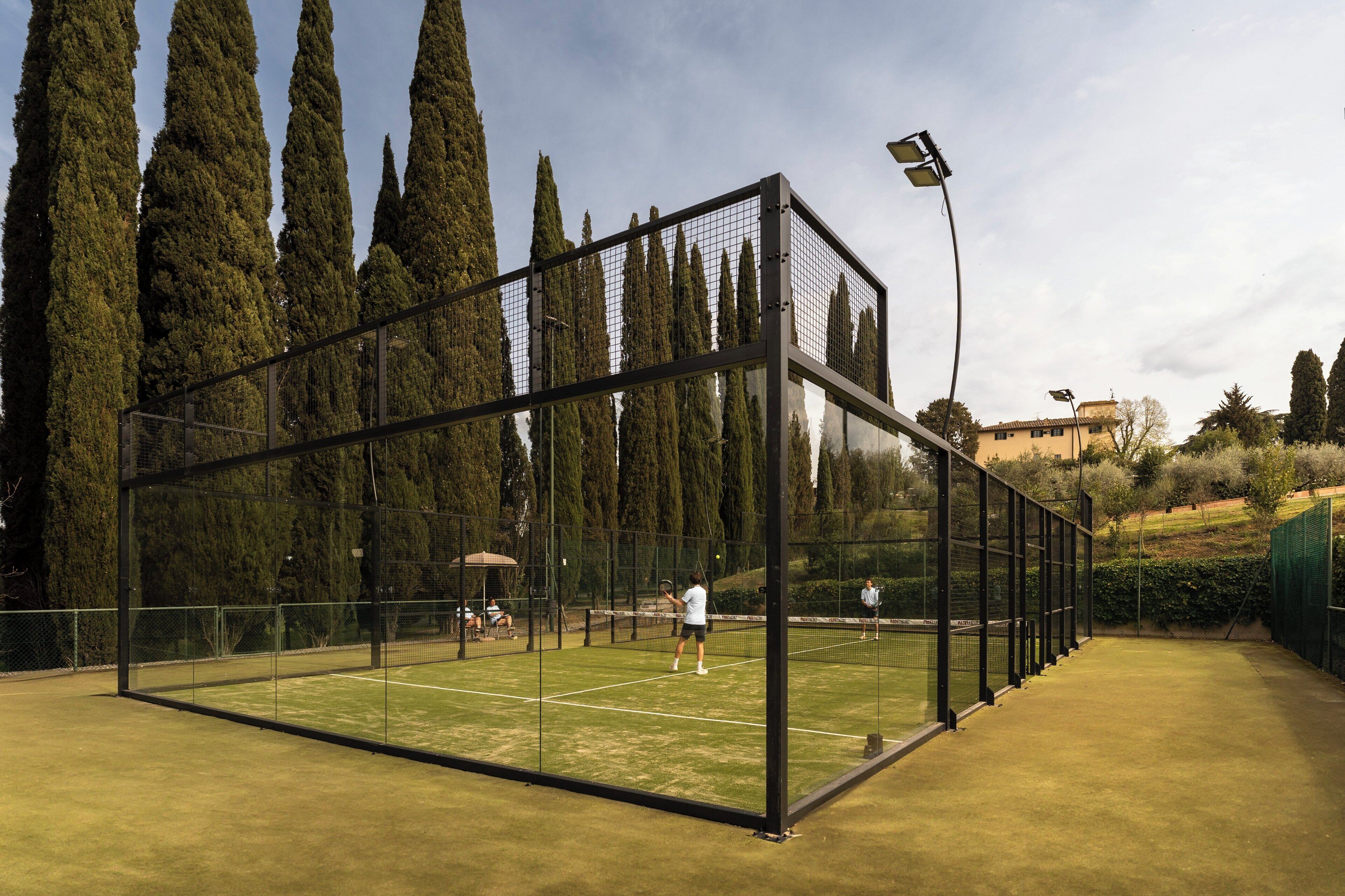Cancha de tenis rodeada por árboles altos y un cielo despejado, con una malla en el centro y una valla de cristal.