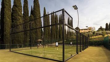 Cancha de tenis rodeada por árboles altos y un cielo despejado, con una malla en el centro y una valla de cristal.