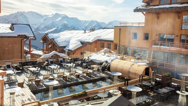A rooftop pool area with wooden deck and lounge chairs, surrounded by buildings with snow-covered roofs and mountains in the background. - Grandes Rousses Hotel & Spa (Huez)