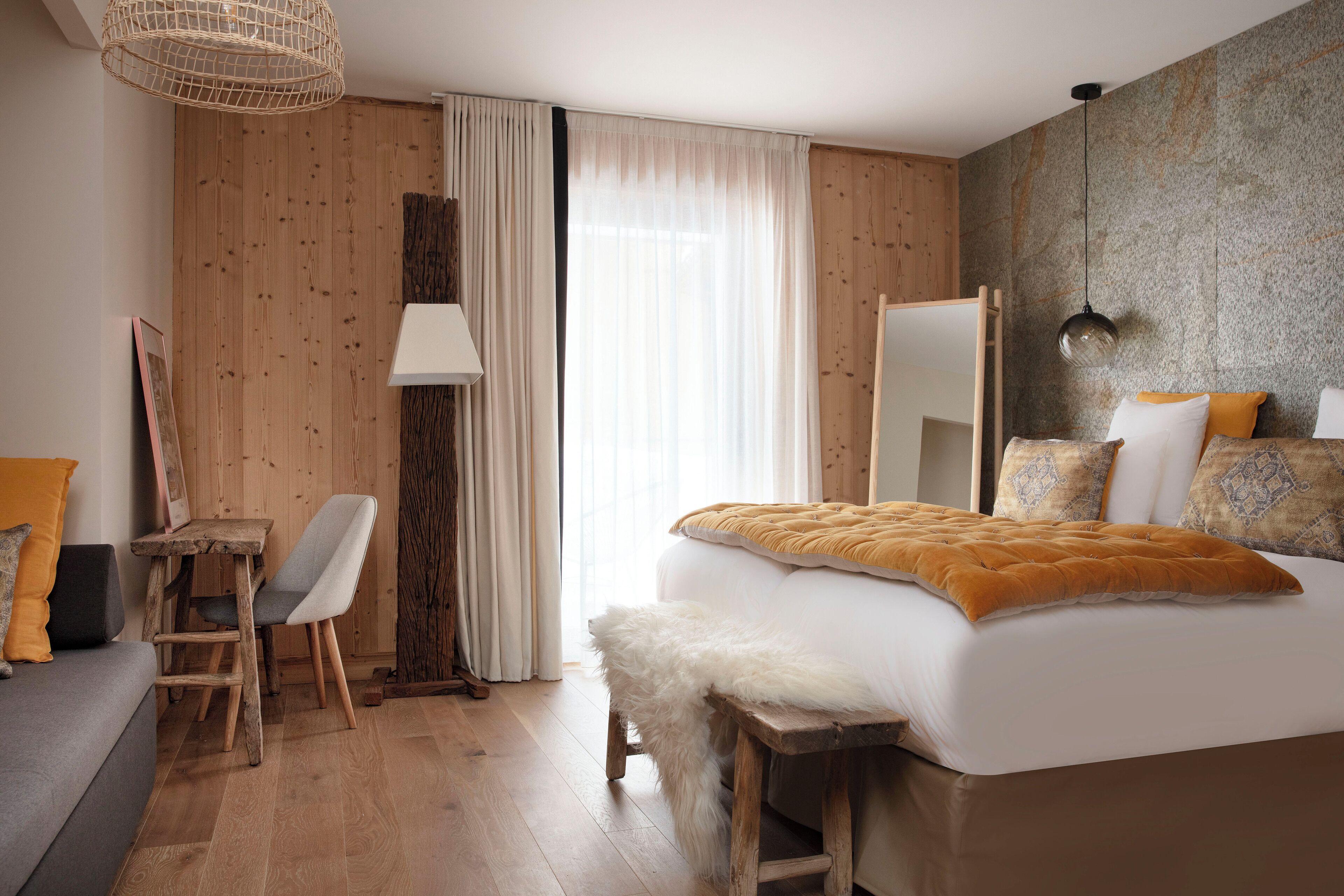 Habitación de hotel moderna con piso de madera, cama grande con un suave plaid blanco, escritorio con silla y una ventana amplia con cortinas traslúcidas.