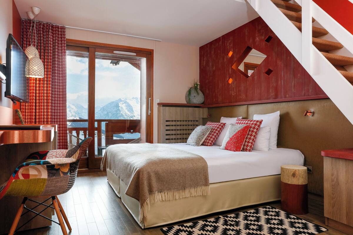 A bedroom with a bed, a desk, a chair, a window with a view of mountains, and a staircase.