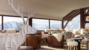 A cozy room with a large window offering a mountain view, furnished with leather sofas, a wooden coffee table, and a decorative antler sculpture. - Grandes Rousses Hotel & Spa (Huez)