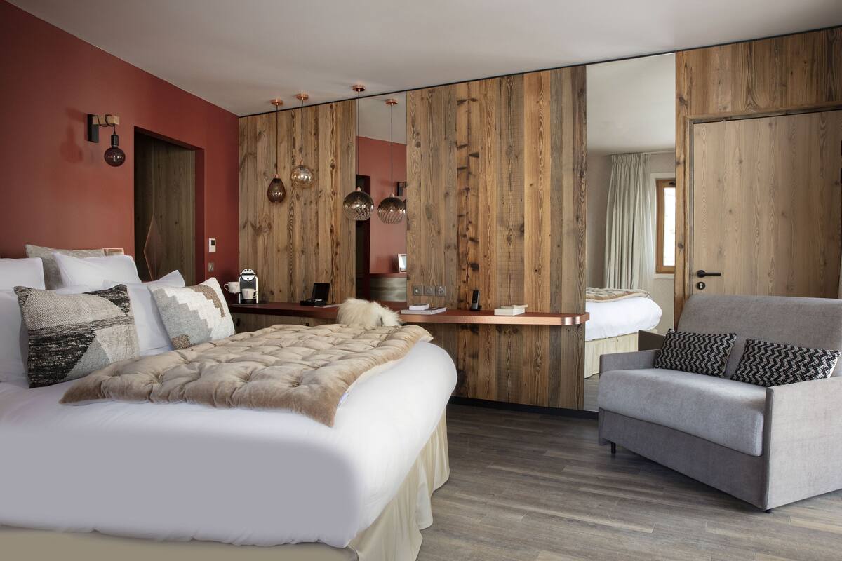 A modern bedroom with a large bed, a wooden accent wall, a desk, and a grey armchair.