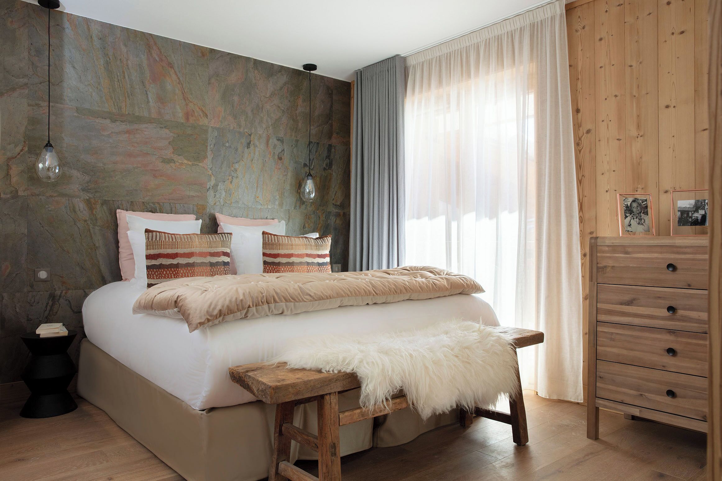 A bedroom with a large bed, wooden headboard, a bench, a dresser, and a stone wall.