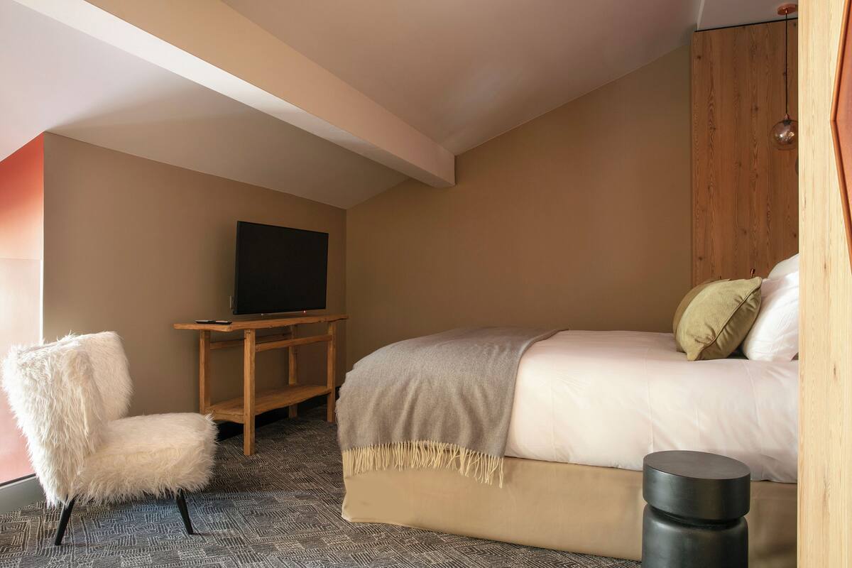 A bedroom with a bed, a chair, a TV on a stand, and a trash bin.