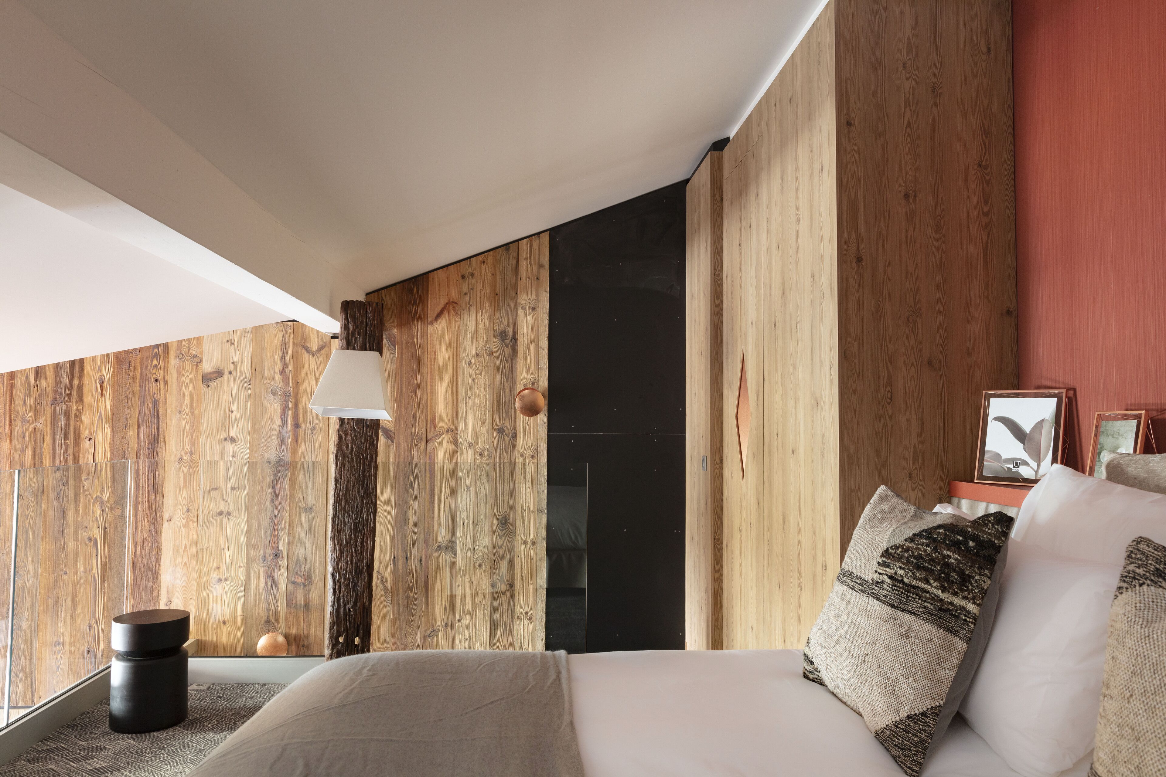 Un dormitorio con pared de madera, una cama con muchos almohadones, una mesita de noche y una lámpara colgante.