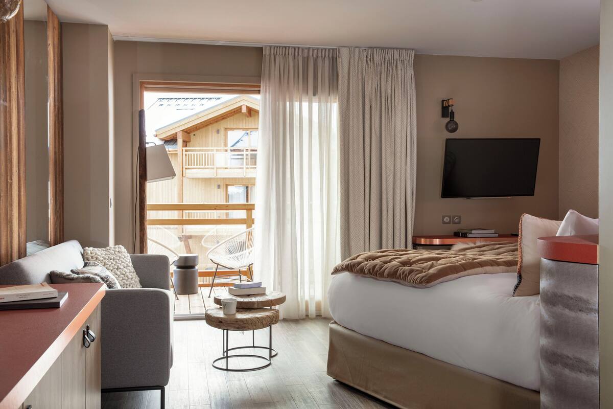 A hotel room with a bed, a desk, a chair, a TV, and a view of a wooden building outside.