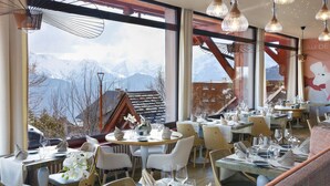A restaurant with tables set for dining, large windows with a mountain view, and hanging pendant lights. - Grandes Rousses Hotel & Spa (Huez)