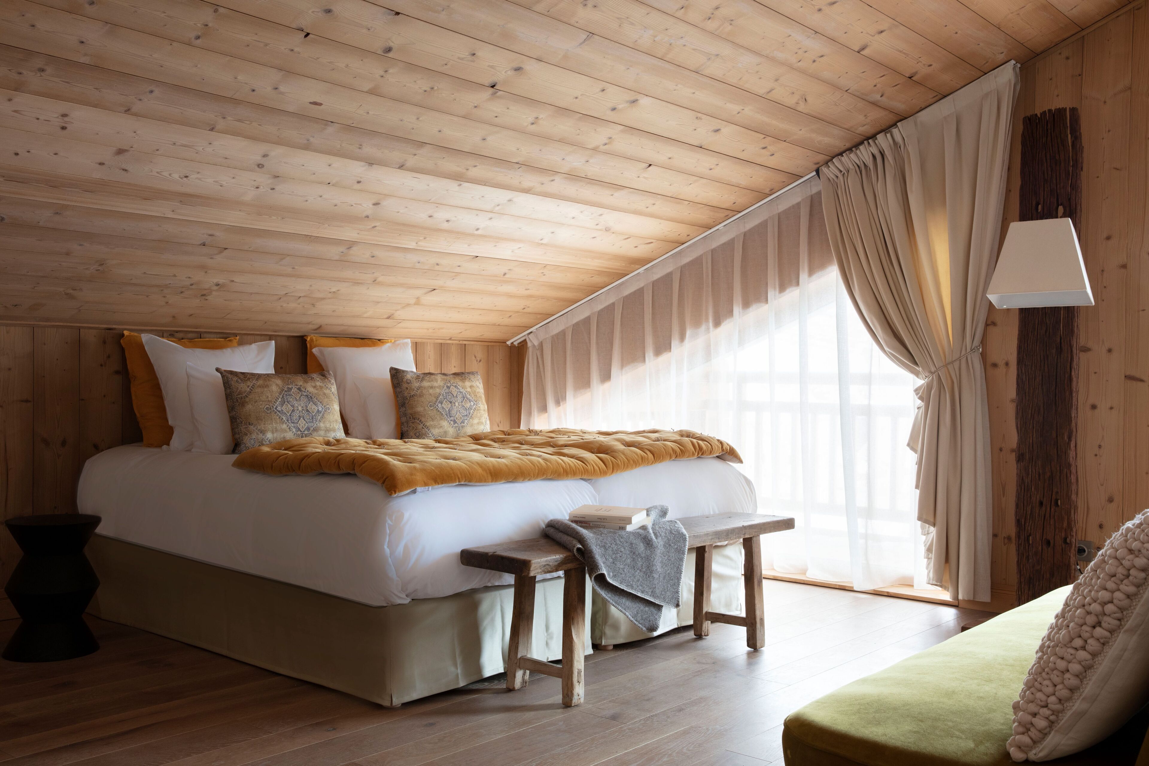 Une chambre avec un plafond en bois, un lit avec une literie blanche et un plaid de couleur moutarde, un banc et une fenêtre ornée de rideaux légers.