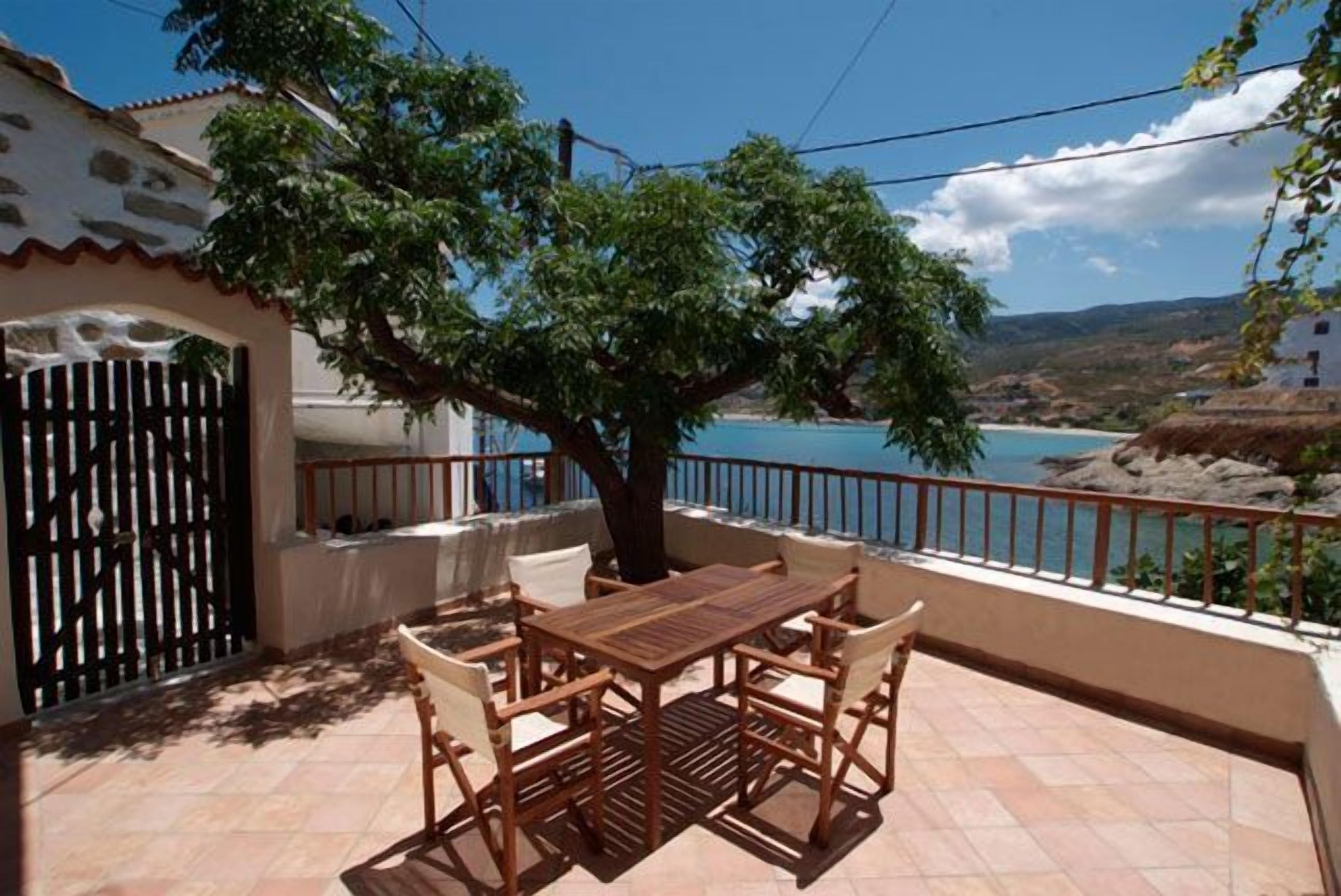 A wooden table and chairs on a balcony with a view of a body of water and a tree.