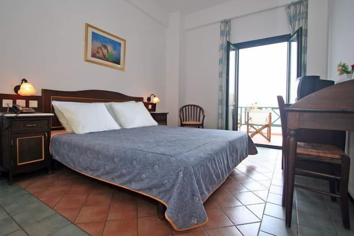 A hotel room with a bed, bedside lamps, a chair, a desk, and a balcony with a view.