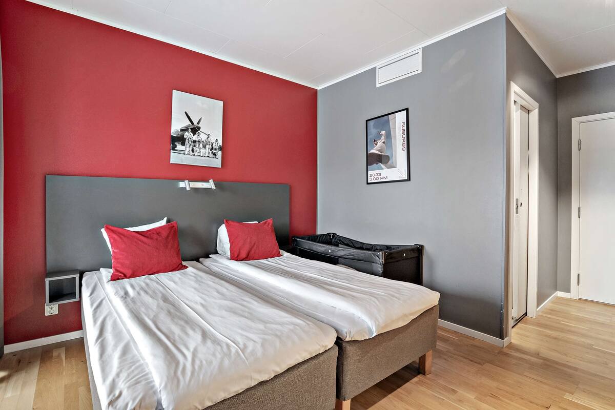 Une chambre d’hôtel moderne avec un grand lit, deux oreillers rouges, une tête de lit grise et une petite table de chevet.