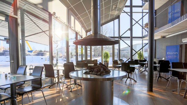 A modern indoor space with a central metallic table surrounded by chairs, large windows offering views of the outside, and a sign indicating the name 'Bonn'. - Aiden by Best Western Stockholm Arlanda Airport (Arlandastad)