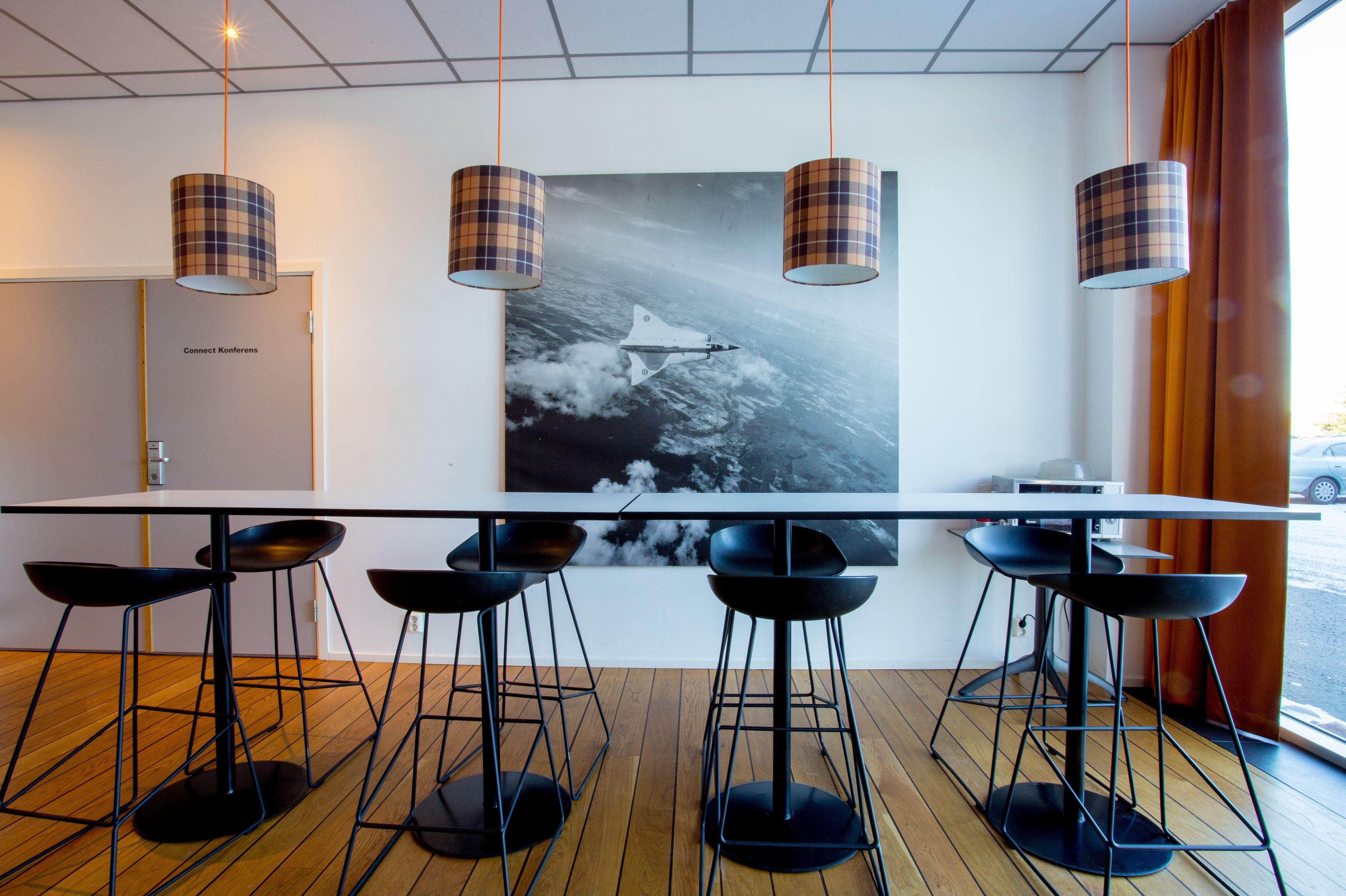A modern conference room with a long table, black bar stools, plaid lamps, and a space shuttle poster on the wall.