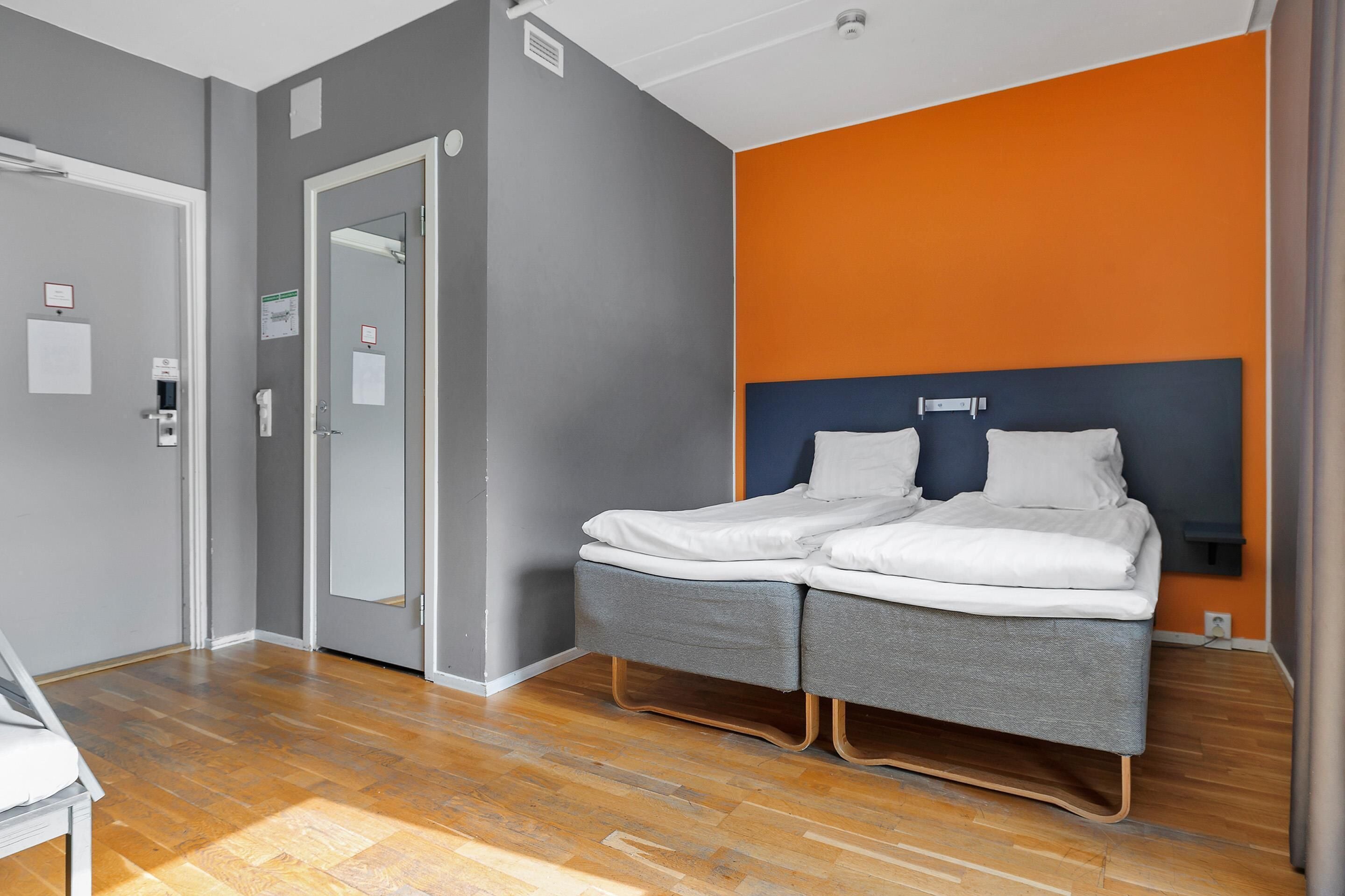 A hotel room with a double bed, a wooden floor, a grey and orange wall, and a white door.