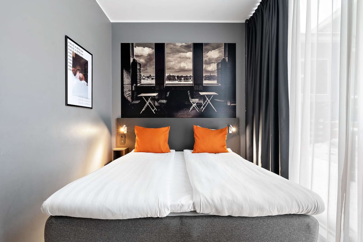 Une chambre d’hôtel moderne avec un grand lit, deux oreillers orange, une vue sur la ville par la fenêtre et un tableau encadré au mur.