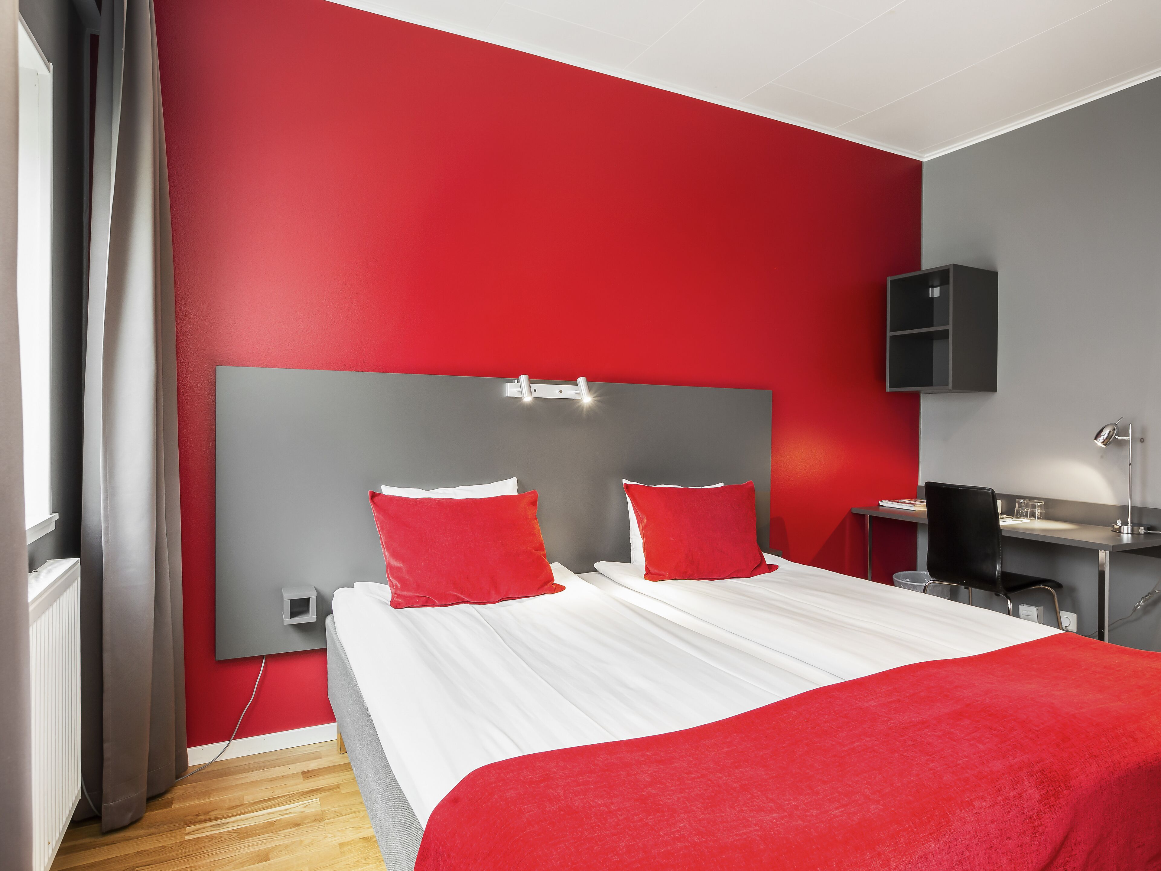 A modern bedroom with a red and grey color scheme, featuring a large bed with red pillows, a bedside table, and a desk with a lamp.