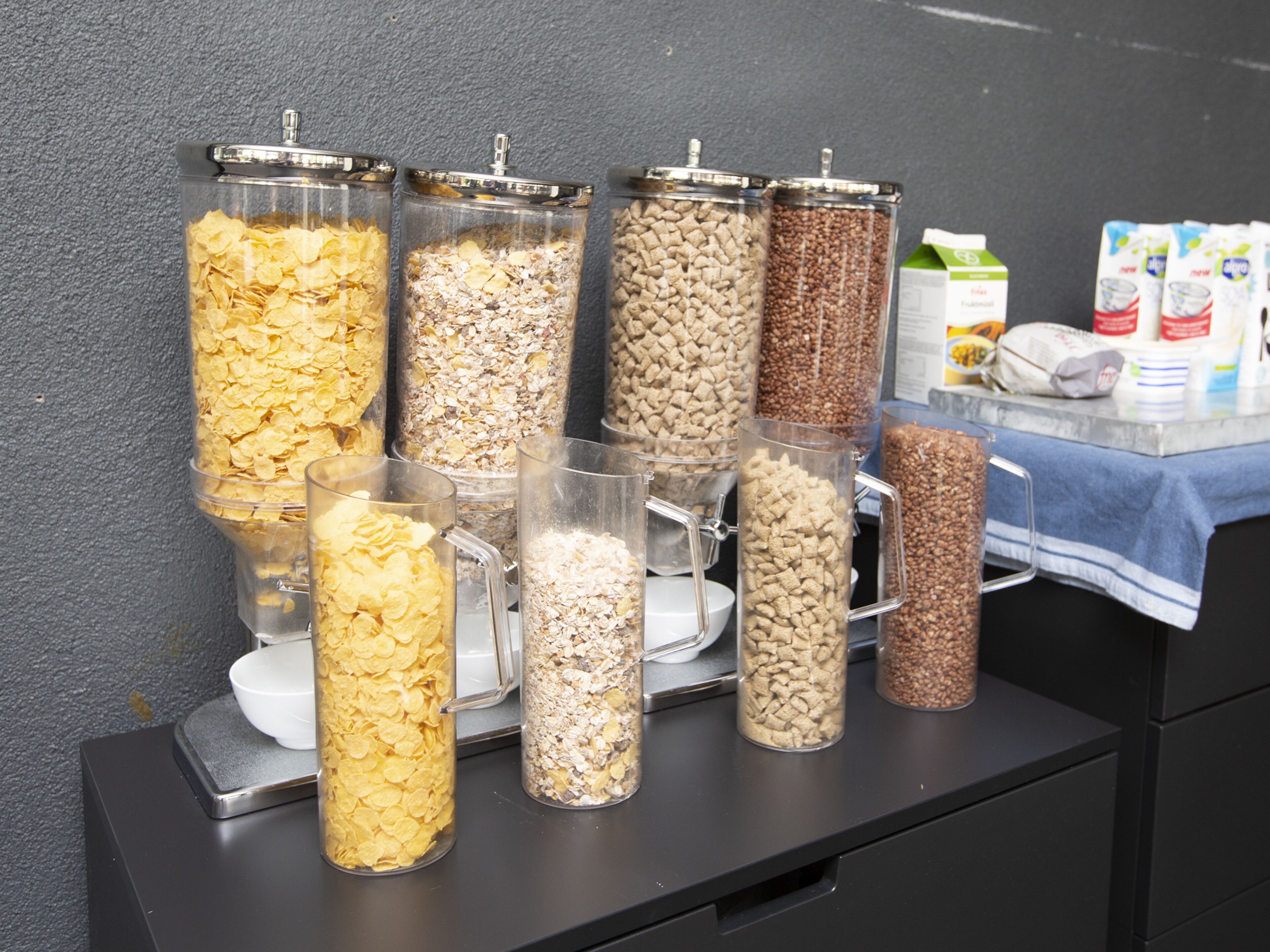Una estación de desayuno autoservicio con diversas opciones de cereales en recipientes transparentes.