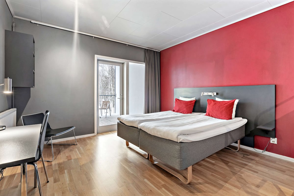Une chambre d’hôtel moderne avec un lit gris, des oreillers rouges, un mur gris et un plancher en bois.