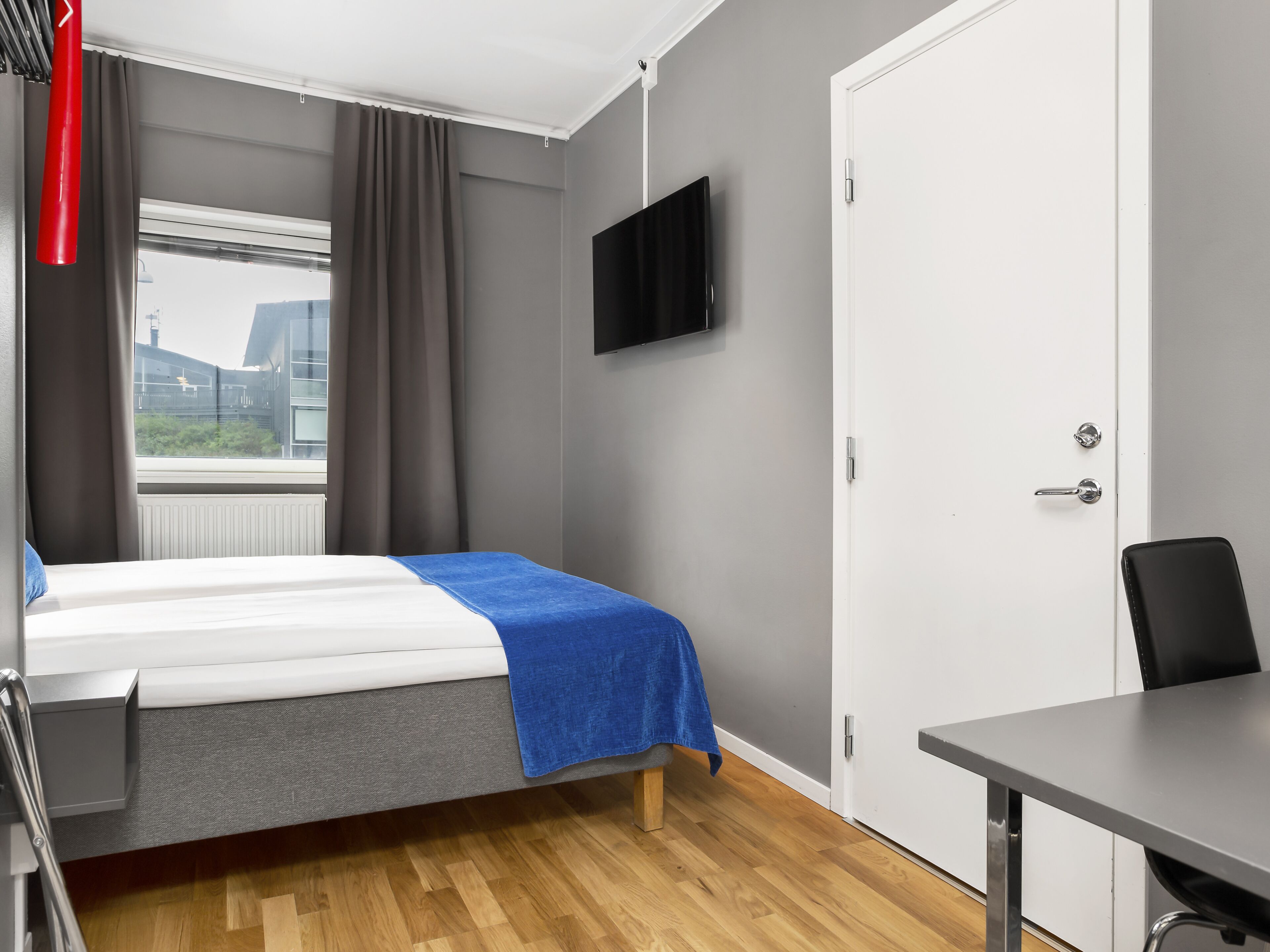 Habitación de hotel con cama, escritorio, silla, televisor y ventana con cortinas.