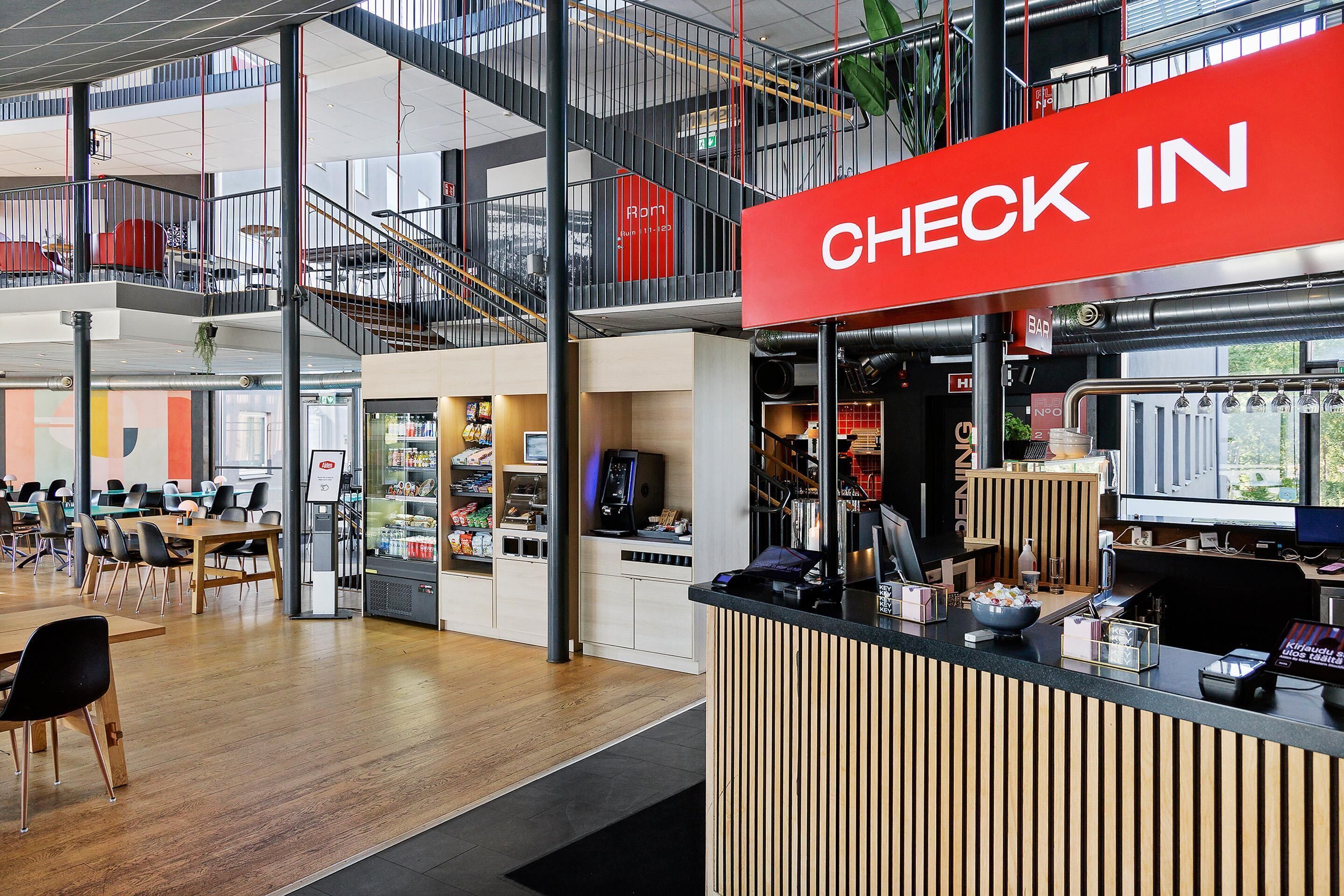 Un interior moderno con un cartel llamativo que dice 'CHECK IN', una máquina de café y un área de descanso con mesas y sillas.